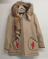 Front view of beige wool coat showing zipper, fur-trimmed hood and appliquéd pockets