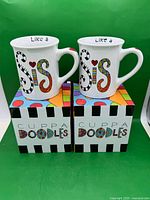 Front view of two Enesco Cuppa Doodles Like a Sis mugs on boxes