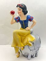 Front view of Snow White figurine sitting on metallic apple holding red apple