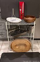 Group shot of entire lot on wire shelves including carving set, platter, potato masher, salad bowl, roasting pan, champagne cooler, carving board, shelves and floor mats