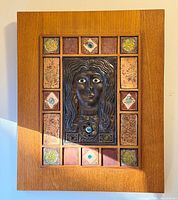 Frontal view of wall plaque showing bronze face, ceramic tile border, glass cabochons, and teak panel