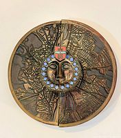 Front view of circular bronze wall sculpture showing map relief, face motif, enamel crest and border