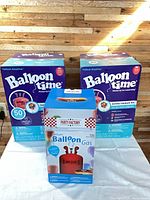 Front view of two sealed Balloon Time jumbo helium kit boxes and one Party Factory helium balloon gas box on table