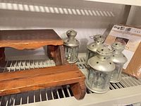 Wooden step stool and four aluminum lanterns on shelf with packaged rug mat partially visible