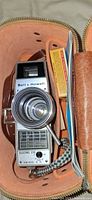 Camera inside tan case showing Bell & Howell branding, lens, solar cell grid, Kodak film box, paperwork