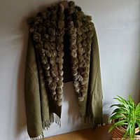 Full view of moss green wool shawl with rabbit pom poms hanging against wall