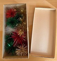 Open cardboard box showing multiple metallic tinsel pom-pom ornaments in various colors