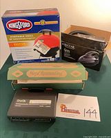 All four boxed items displayed: Kingsford grill box, Majestic Bells wind chime box, Riccar Gem vacuum box, OneClik flashlight kit case