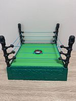 Overall view of WWE toy wrestling ring showing green mat, black posts, blue ropes