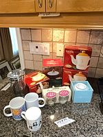 Group shot of all items on countertop: boxed Tim Hortons teapots, loose teapot, mugs, shaker, jars, blue box