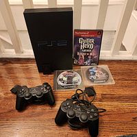 PS2 console standing with two controllers, Guitar Hero Encore case and two loose discs