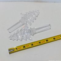 Two clear crystal bottle stoppers shaped like Christmas trees beside ruler showing about 5 in length