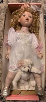 Full view of porcelain doll with dress, shoes and teddy bear inside box