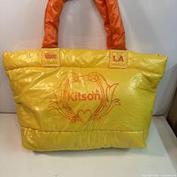Front view showing yellow puffer tote, orange handles, logo patches