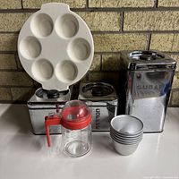 Front view of all items grouped: three chrome canisters, egg dish, glass syrup dispenser, stack of pudding moulds