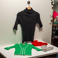 overview of all items arranged on table: black turtleneck hanging, green cardigan, red booties, grey gloves