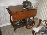 Solid wood tea cart on wheels with folding leaves closed, showing upper surface and silver serving pieces on top and bottom shelves.