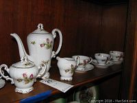 Overall view of tea pot, sugar bowl, cream pitcher, and several cups with saucers on wooden shelf