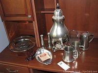 Full display of pewter coffee urn with lid and warmer, two open mugs, lidded mug, tray, bud vases, three wooden plate stands, and glass ship mug arranged on wood cabinet.