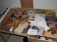Top-down photo showing woodworking books, carving tools, paint brushes, gloves, and small hand tools laid out on a table with a 3D kitchen scene relief in wood.