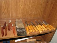 Full view of the 12 wood carving tools arranged on a wooden shelf alongside the AMT fabric pouch and a measuring ruler.