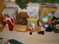 Boxes containing assorted Christmas wreaths, a red nylon angel fabric decoration, and a wooden nutcracker figurine along with a large colorful snowman lantern figure holding a broom on floor.