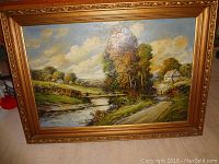 Full view of the oil on canvas landscape painting in ornate gold frame showing the entire scene of river, trees, small houses and road.