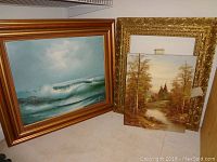 Frontal view showing both oil paintings; the seascape in wood frame and the landscape detached from its gold ornate frame.