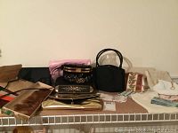 View of multiple handbags and clutches including black beaded, floral embroidered clutch, metallic gold clutch, and pink handbag with a box of natural hair pieces.
