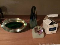 Eye-shaped piece with green and amber iridescent finish next to dark blue pyramid piece and small colorful paperweight in box on wood surface
