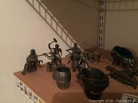 Photo of six pewter figurines including men in various poses and attire displayed on wooden shelf; also shows a metal bucket and cast iron shoe pincushion