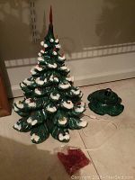 Full view of the ceramic Christmas tree with white doves and base on tile floor, showing wired cord for lights and red star topper near the tree.