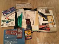 Photo showing a variety of calligraphy and art supplies including watercolor paper, watercolor paints, calligraphy pen nibs, and books.