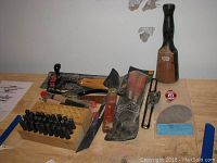 Photo showing full lot of woodworking tools including small hand plane, stamping letters in wooden block, carving tools with cork handles, large wooden handled chisel, hand saw blade and other small tools.