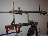 View of multiple pipe clamps hanging on a rack with some showing orange jaws and yellow end caps, as well as a black tool belt below them.