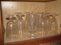 Twelve clear crystal goblets with etched floral designs on a wood shelf, some upside down showing the engraving details.