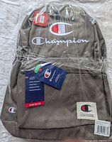 Front view of Champion grey backpack in plastic wrap, showing logos, tag, and front pocket