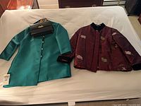 Overview of all three items on bed: teal Bailey jacket, maroon embroidered jacket, black D.W.L purse