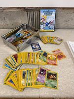 Metal tin open with stack and spread of Pokémon cards, DVD case in background