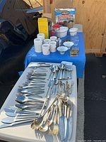 Overview of lot showing flatware laid out and secondary table with mugs, bowls, small glasses, candles and Kirkland trash bags