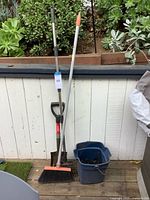 All four items leaning against wall: shovel, broom, rake, bucket