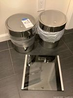 Two stainless steel waste baskets and glass bathroom scale on tiled floor
