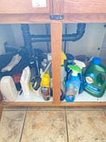 Full view of under-sink cabinet showing cleaning products, Lysol jug, spray bottles, garbage can, textiles