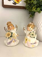 Front view of both Lefton birthday angel figurines showing month banners and flowers