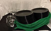 Stack of black plastic plates in green cloth with five clear plastic stemmed wine cups and clear punch ladle