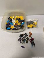 Plastic tub with assorted coloured Mega Bloks bricks, separate bag of yellow bricks, figures nearby