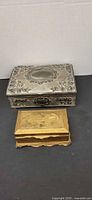 Two metal jewelry boxes, larger silver-tone with floral repousse and smaller gold-tone with embossed lid
