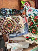 Overview of assorted embroidered textiles stacked, showing cross-stitch panels, cushion covers and beaded sash