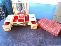 Jewelry box open with red velvet interior, side drawers extended, floral sleeve beside