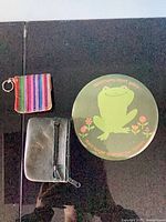 All three items laid out: round frog tin, black coin pouch, woven striped pouch